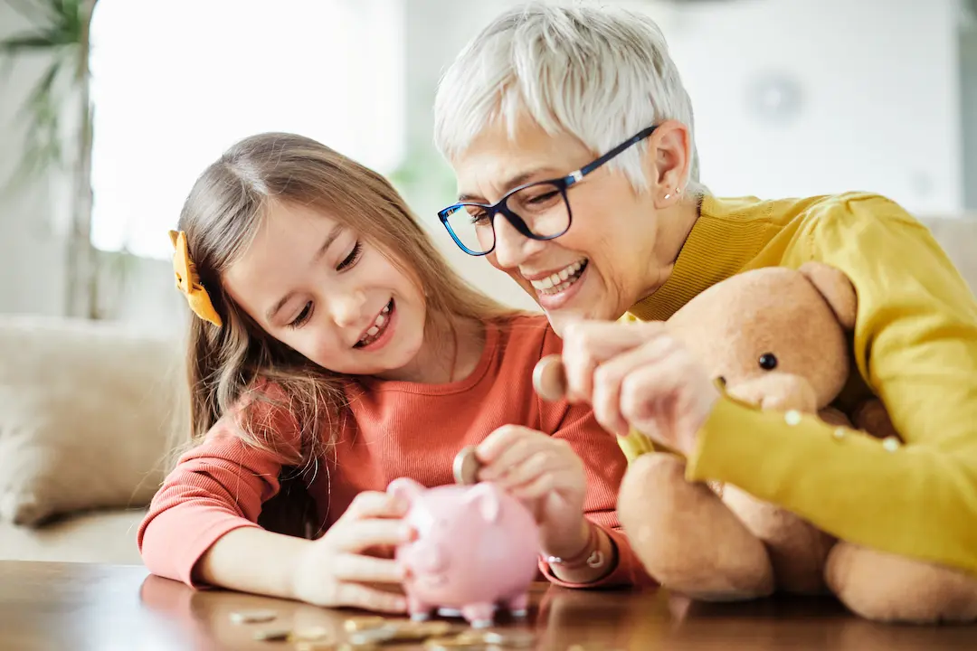 Grandmother and granddaughter smiling while saving coins in a piggy bank together at home. 